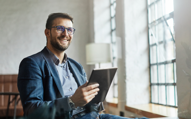 Foto de un hombre con barba y gafas de marco oscuros, vestido con camisa y saco. Tiene una tableta en sus manos conociendo información de los términos banca de inversión y mira hacia el horizonte sonriendo.