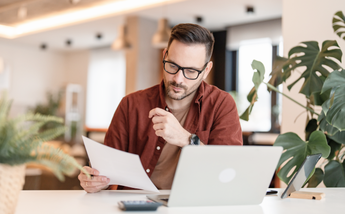 Foto de un hombre con gafas y barba ubicado en la mesa de su oficina mirando los documentos legales Banca de Inversión y con su computador portátil.
