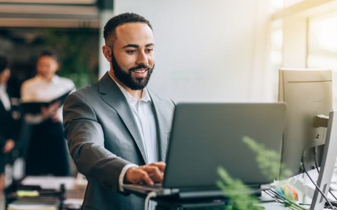 Foto de un hombre joven barbado con traje gris sonriendo y consultado en un computador portátil la información sobre protección de datos Banca de Inversión.
