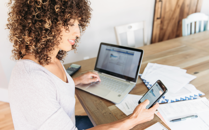 Información financiera Banca de Inversión Foto de una mujer de cabello rizado color castaño, mirando la información financiera Banca de Inversión en documentos, su celular y computador portátil.